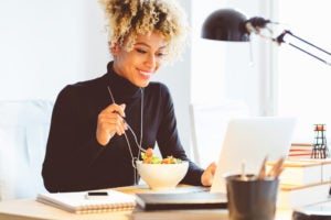 Black woman sitting at the desk in an office and eating lunch, using laptop at the same time.