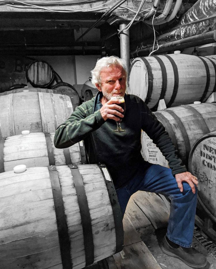 An older man sitting among beer barrels with a beer in hand.