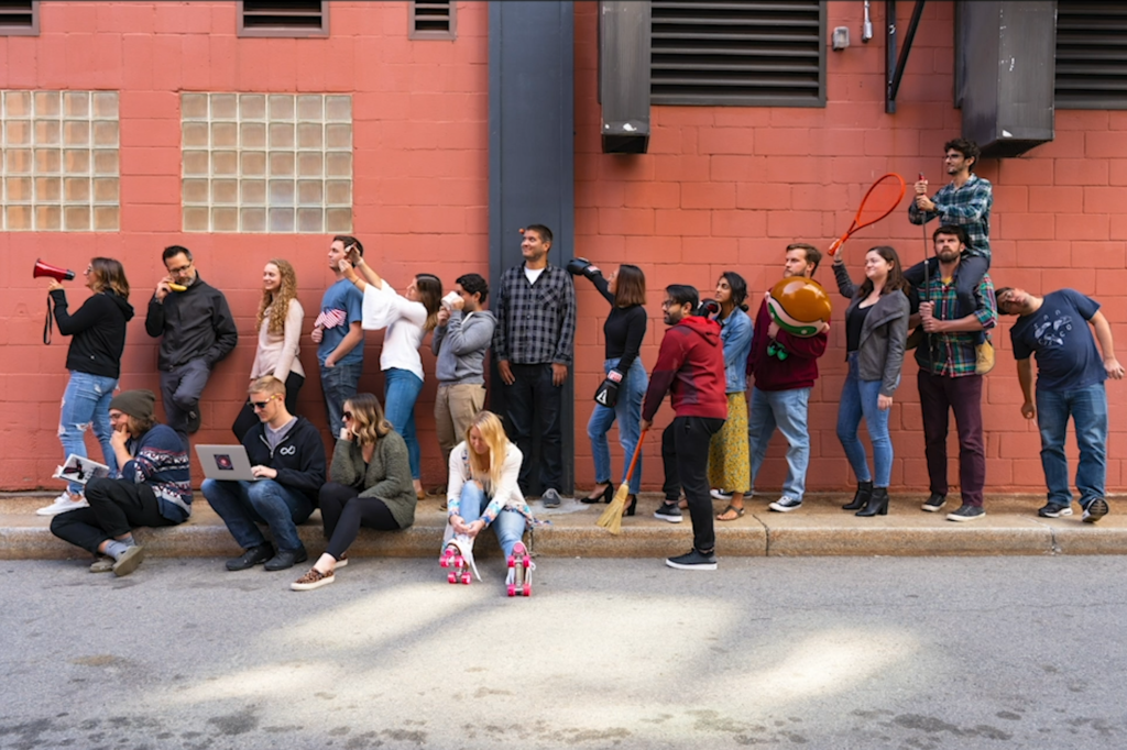 A group standing beside a red brick wall, a few people sitting on the ground in front of them, one person wearing rollerskates