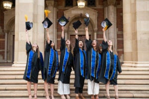 Six college graduates wearing their cap and gowns celebrate with a photo shoot in front of the library. They hold their caps high in the air as they prepare to throw them simultaneously.