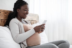 Black Pregnant woman Spending Time With Smartphone At Home, sitting on bed