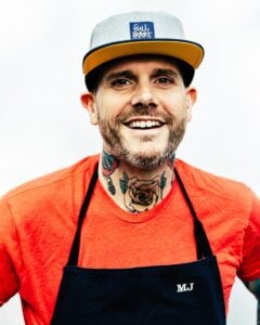 Matt Jennings in an orange shirt and black apron, wearing a baseball hat with the full heart logo on it. He has tattoos of roses on his neck.