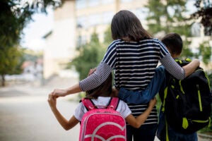 A family is walking to school together on a sunny day. The mother is in the middle with both arms over her children, who are embracing her.