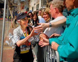 Benoit Samuelson, now an older adult with gray hair in a ponytail underneath a navy baseball cap greets a large group of her fans who are gathered behind a metal barrier. 