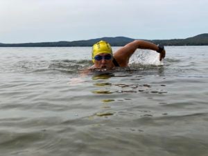 Patricia Gallant-Charette — a stately, middle-aged woman wearing a yellow bathing cap and blue goggles — pops her head out of the water mid-stroke and takes in a breath of air as she swims across the English Channel. The scene is set against a strip of blue-green mountains in the background.