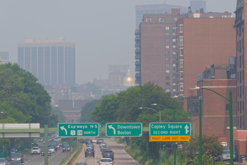 The golden dome of the Massachusetts State House and surrounding structures are partially obscured by the thick smoke. Cars drive down the highway towards the city.