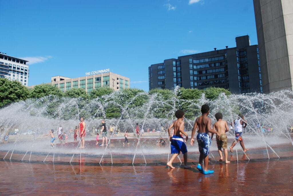 Children cool off in the Reflecting Pool at the Christian Science Plaza during a heatwave. They run towards jets of water spraying in a circle.