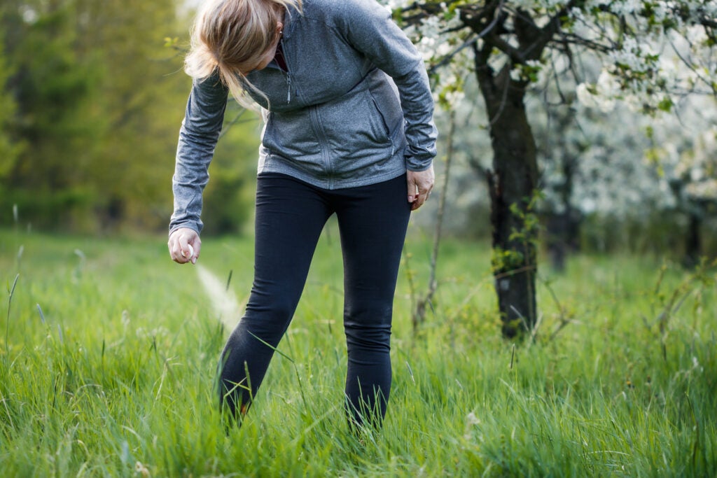 A woman wearing sports leggings and a gray zip-up sprays insect repellent against her leg. She wears her blonde hair in a pony tail, and stands in a bright green field.