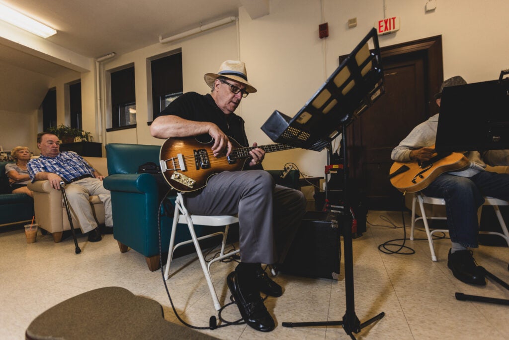 An older adult man plays the electric guitar sitting down in front of a music stand in a senior living community. 