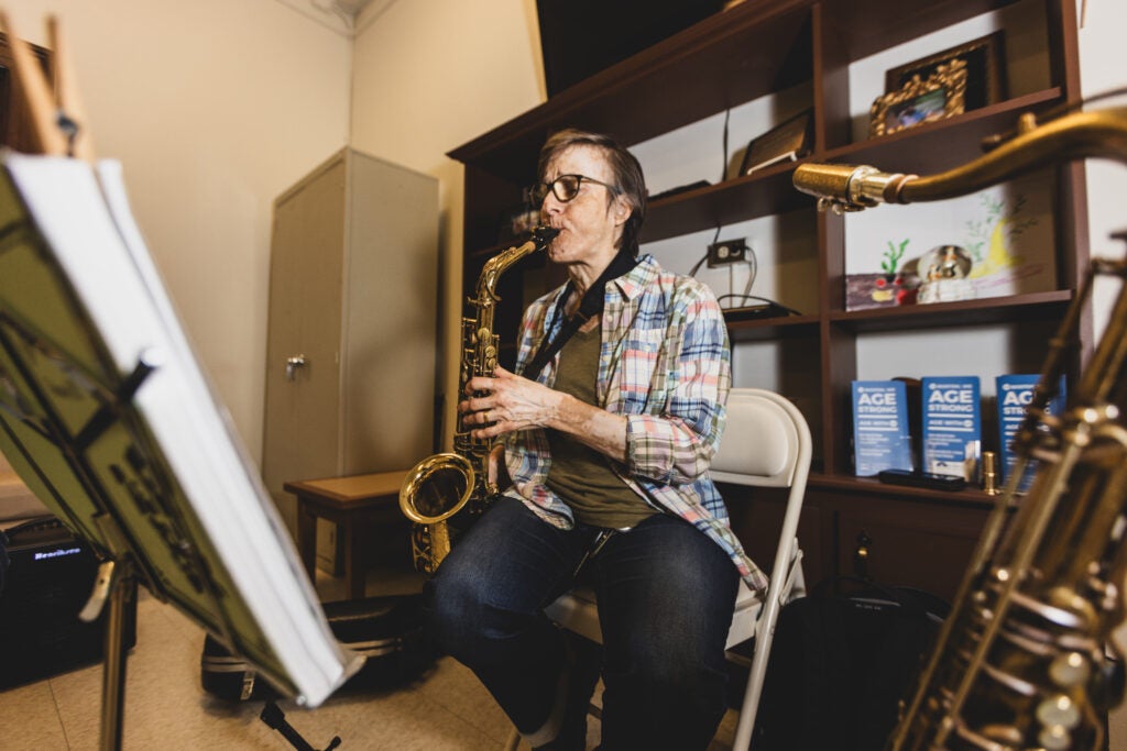 An older adult sits on a chair in front of a music stand playing the saxophone.