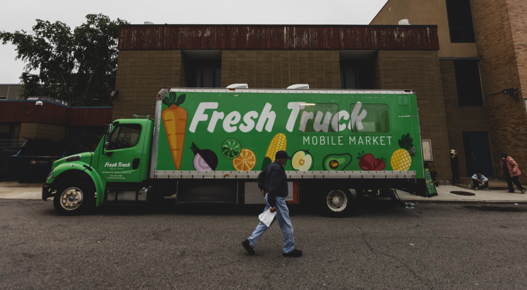 A person walks alongside the profile of the Fresh Truck, which bright green exterior lights up a gloomy Boston day.