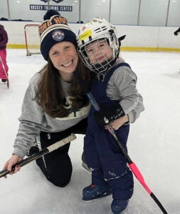 A mother wearing a beanie and sweatshirt poses with her daughter on the ice during hockey practice.