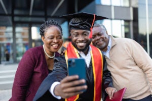 A man weating a graduation cap and gown takes a selfie with a well dressed older couple behind him.