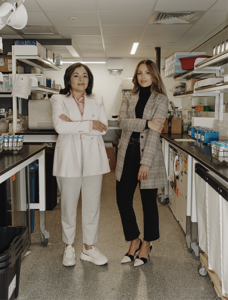 Two women in business clothes standing with arms crossed in a lab.