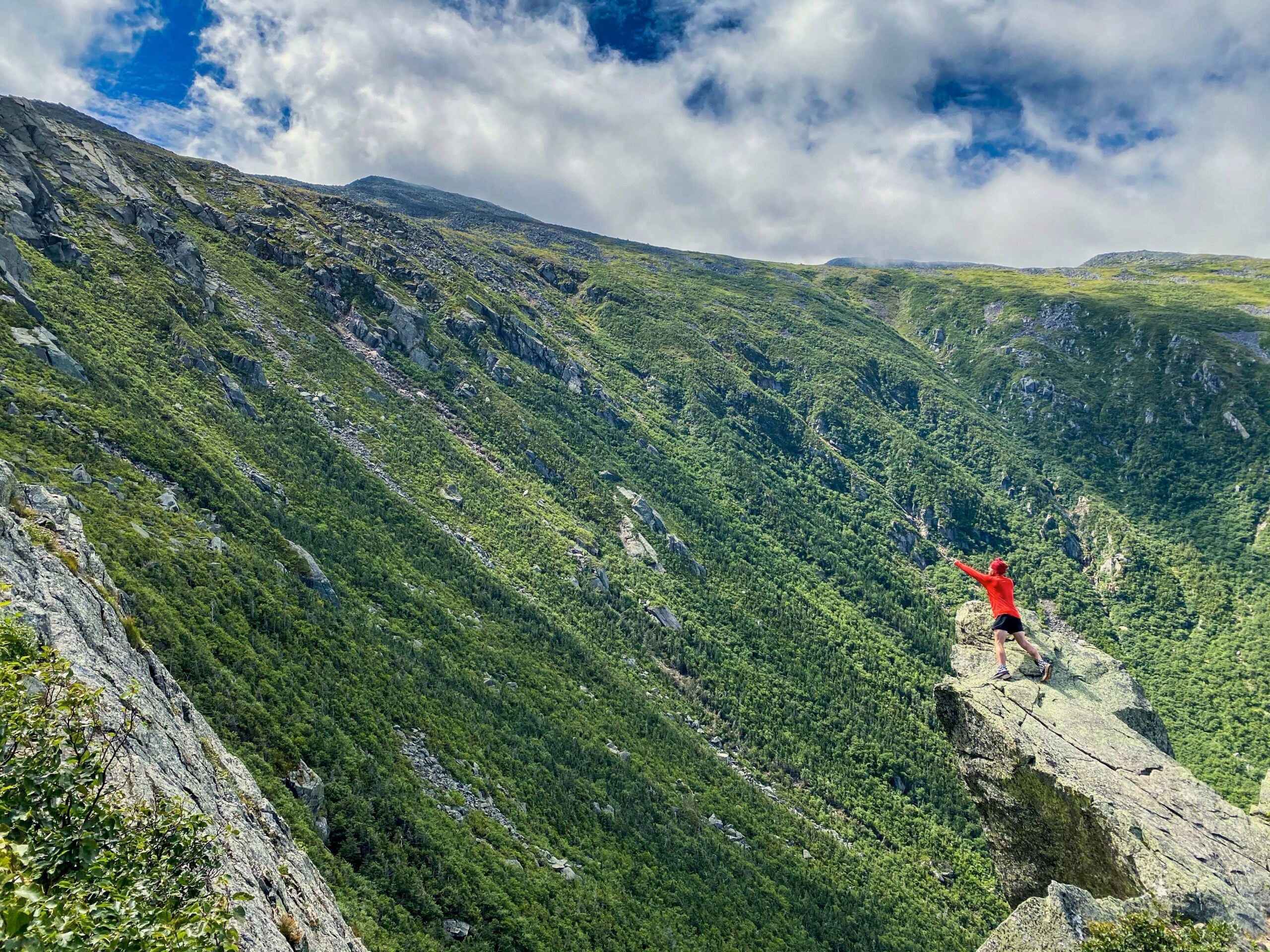 A hiker wearing a red hoodie stand on the edge of a cliff reaching out towards another mountain.