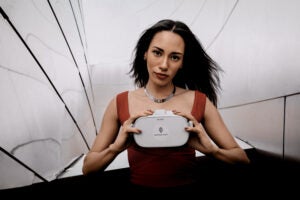 Woman with dark hair and red shirt holding virtual reality headset against white background.