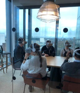 Group of people sitting at conference table wearing virtual reality headsets in room with glass walls overlooking the city.