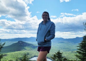 A woman in a blue hoodie stands in front of a scenic landscape.