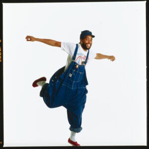 Man in baggy overalls posing standing on one leg with arms outstretched..