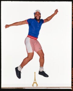 Man in active wear poses jumping above a small model of the Eiffel Tower with arms outstretched.