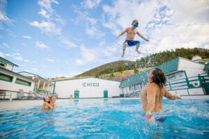 A young boy is in mid air after being thrown up and out of a man with his back to camera. A woman and girl also in the pool, watch from the background.
