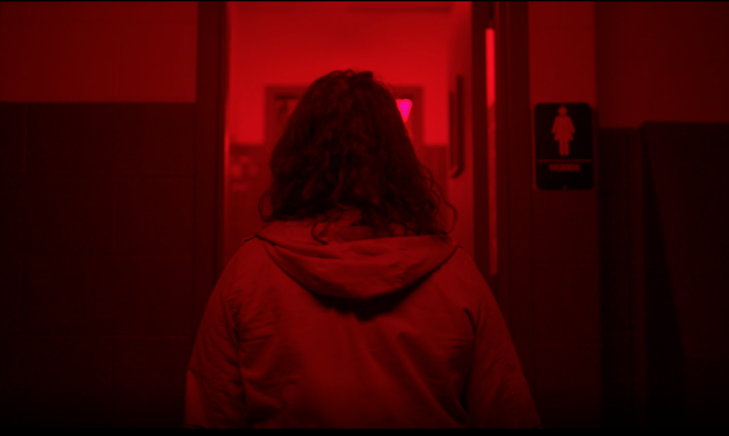 Under red lighting, a woman facing away from camera walks towards a public women's bathroom.