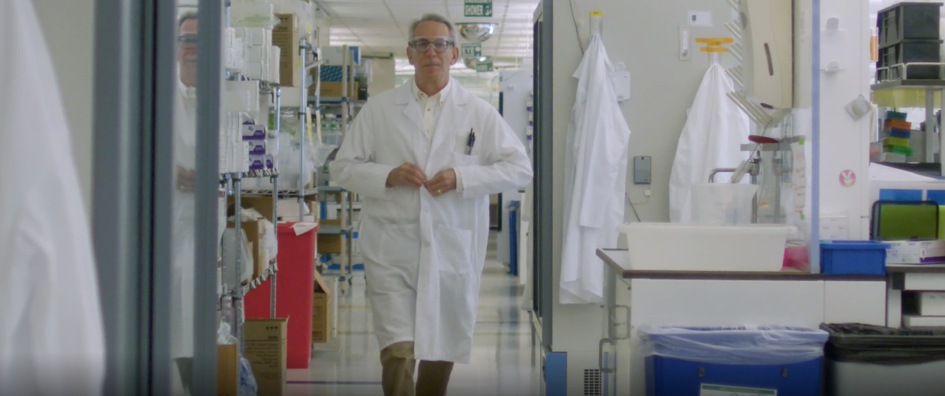 In a lab setting, an older man with white hair and glasses buttons up a lab coat as he walks towards the camera. 