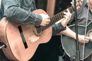 Close shot of two men's hands playing two wood guitars, the men strumming wear a jean jacket and blue shirt respectively.