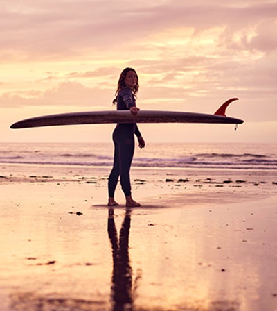 A woman in a wet suit walks across a pink and yellow-lit sunset beach while holding a surf board. Her shadowy reflection gleams in the wet sand in front of her.