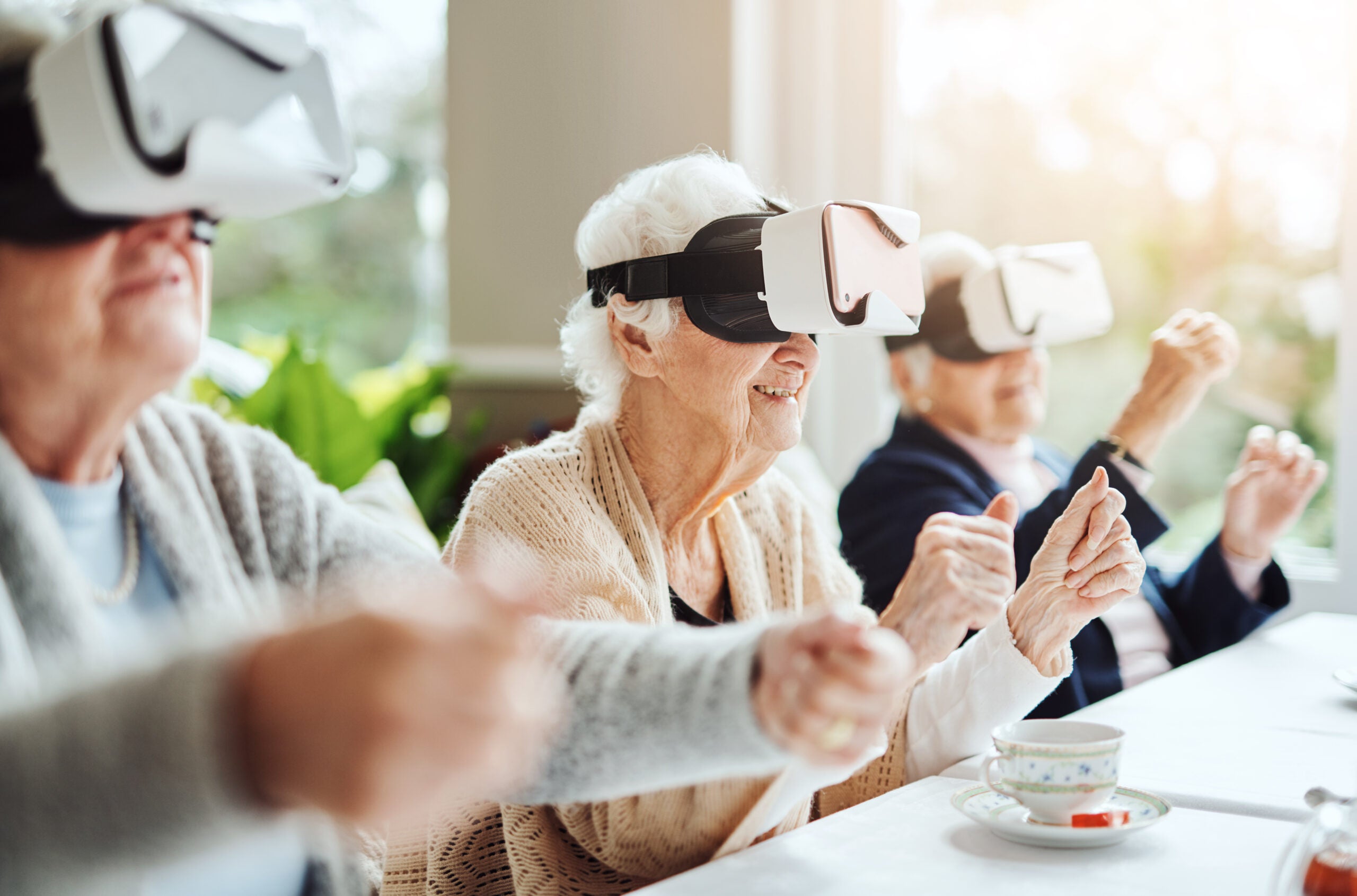 Senior citizens engaging in virtual reality while wearing VR headsets and sitting at a long conference table. Their arms are positioned as though they're driving a car. 