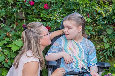 A child with down syndrome wears a sky blue, pink and yellow T-shirt and her dark blond hair in braids. She turns her head to listen to a social worker who has knelt down to talk to her.