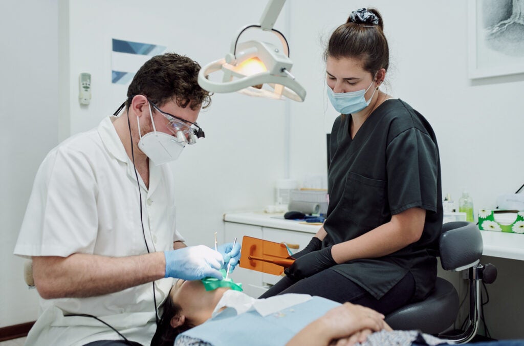 A dental specialist wearing a white button-up shirt and protective gear conducts a cleaning on a woman laying down. A dental assistant in black scrubs and a medical mask sits nearby holding dental appliances.