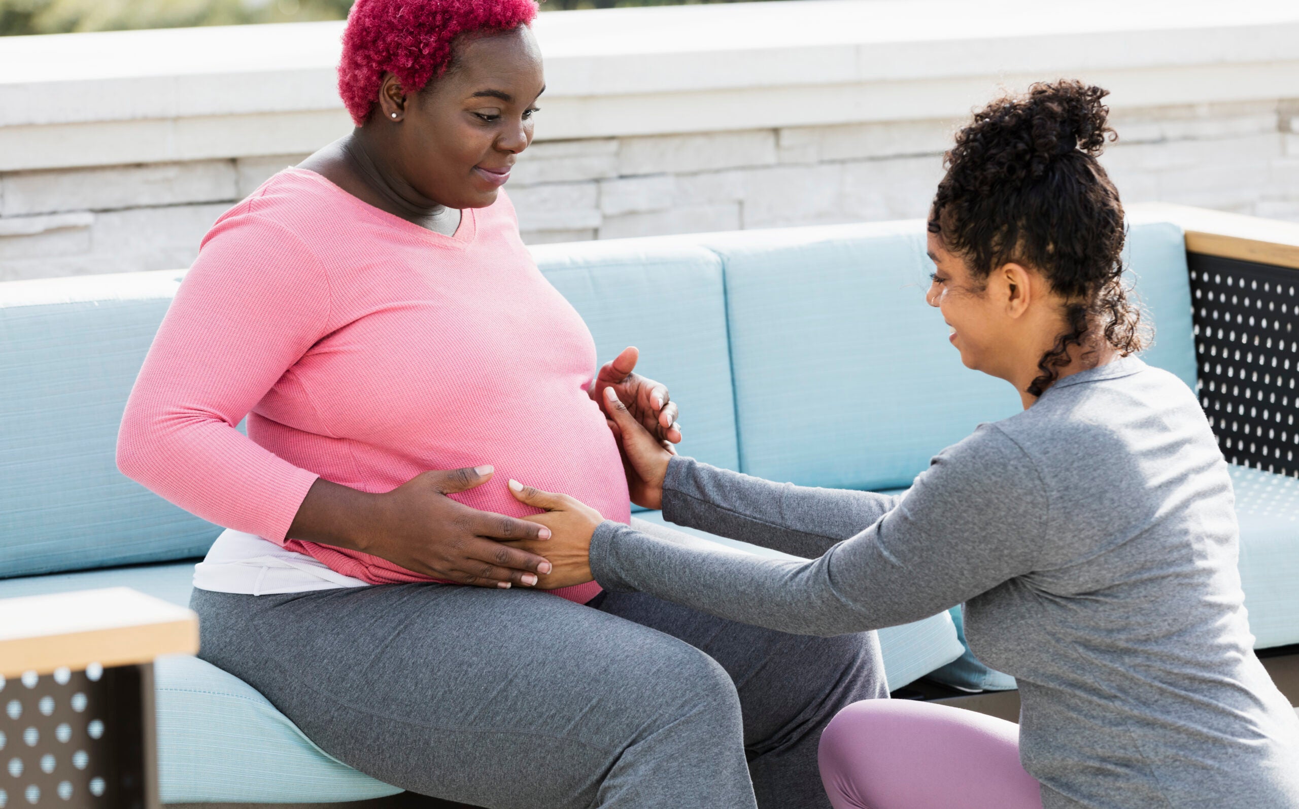 Pregnant woman with short, bright red dyed hair wearing a long-sleeved pink shirt and grey yoga pants sits on a sky blue couch as a doula touches her stomach.
