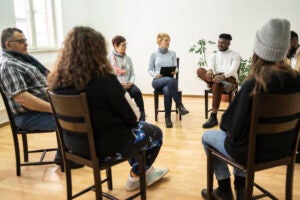 A diverse group sits in a circle of chairs, listening to each other in a group counseling setting.
