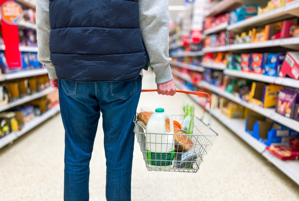 A man wearing jeans and a navy blue down vest over a grey sweatshirt stands in the middle of a grocery store aisle with a shopping basket full of milk, bread, and other foods. 