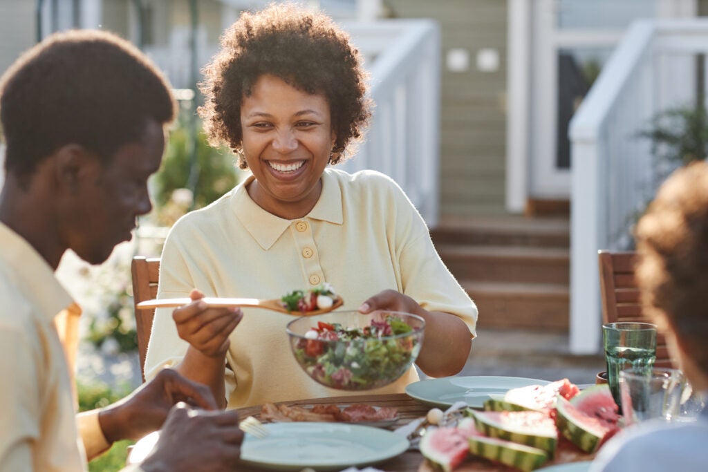 A woman sits at a table outdoors with her family, wearing yellow button-up shirts. The woman smiles at a young buy while serving him a leafy, green salad.