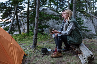 Blond woman and brunette man sitting on a log in the woods holding coffee mugs and looking out into the distance contentedly. They're in the woods in front of a boulder. An orange tent is in front of them
