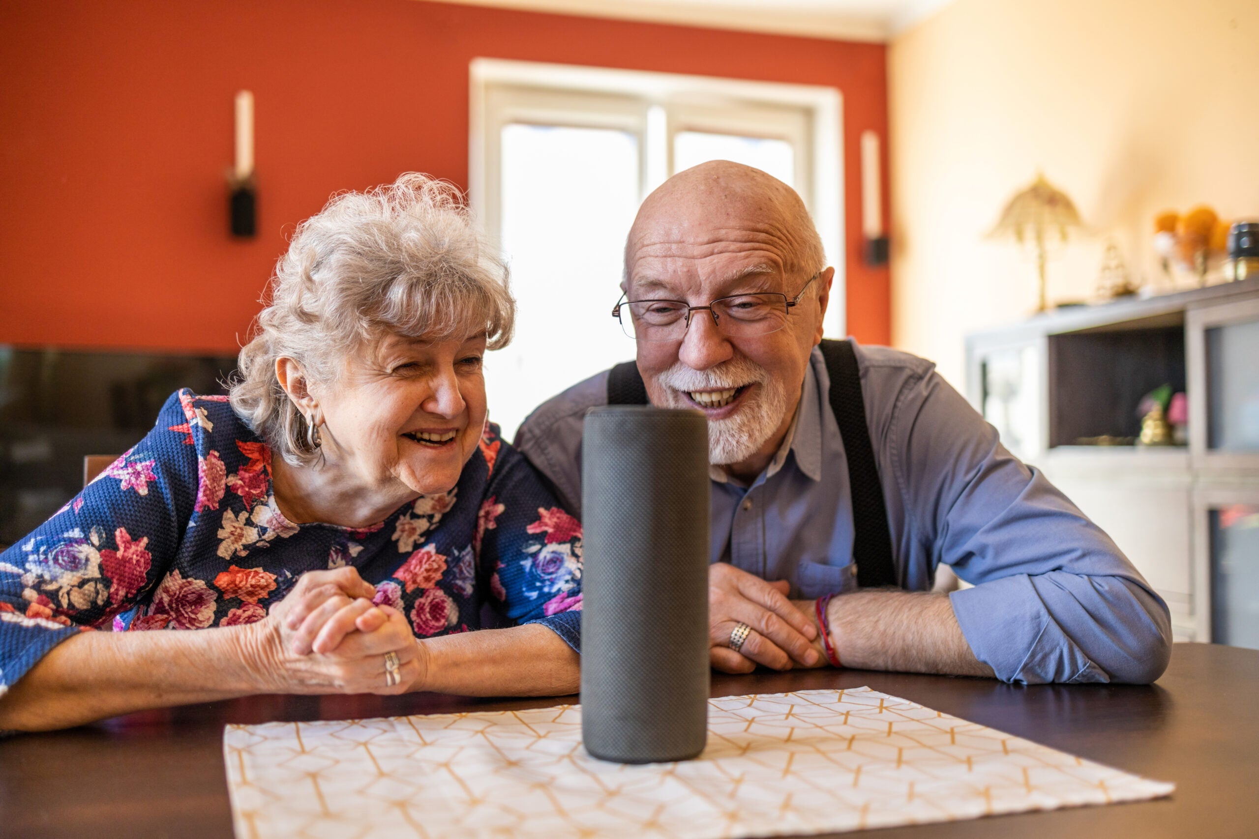 Elderly hetero couple excitedly use a voice-enabled virtual assistant speaker at home.