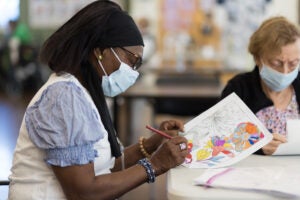 A Black woman wearing a mask sits at a table doing adult coloring