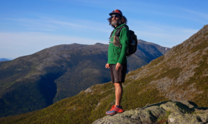A man with a full beard and long hair in athletic clothes with a backpack stands on a rock in front of a mountain