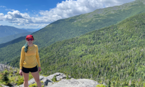 A woman in athletic gear and a red hat stands on the edge of a mountain overlooking a sea of trees