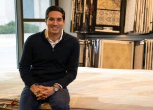 A man sits on a pile of rugs with his hands in his lap smiling at the camera.