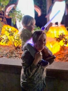 A young woman carries a small boy with a toy lightsaber on her shoulders as they walk through a light show at night