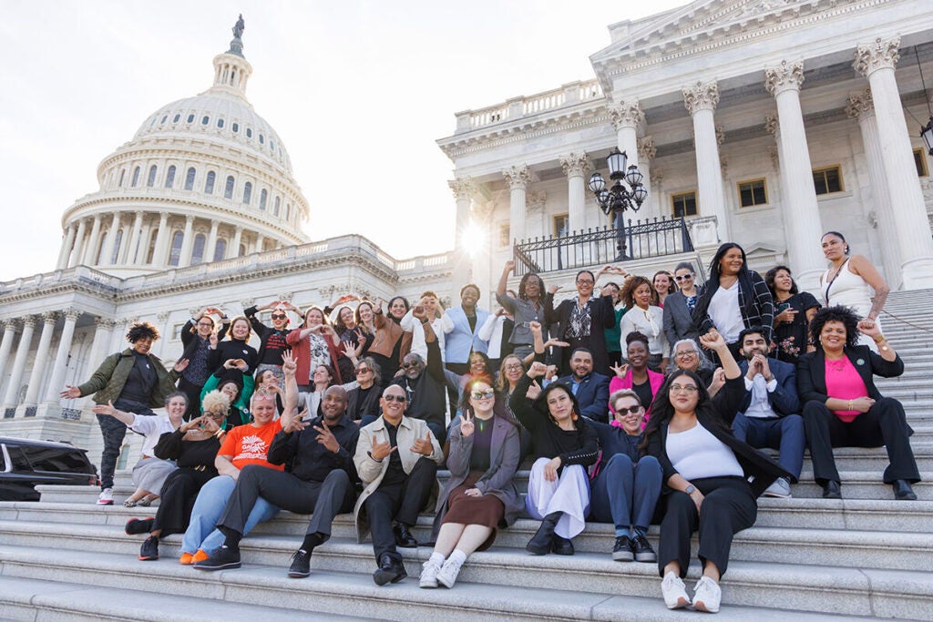 A group of 30-40 community health workers sit on the steps to Capitol Hill, smiling and pumping their fists in the air.