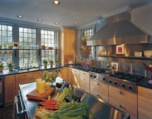 Large kitchen with colorful vegetables on a center island, wood cabinets, metal fixtures, and a wall of windows.