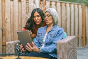 A senior woman sits on outdoor furniture holding a tablet as a young woman looks over her shoulder.