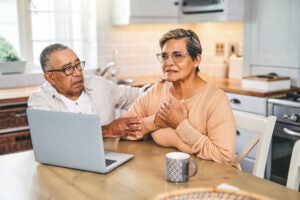An elderly man and woman sit at a kitchen table in front of a laptop looking concerned.