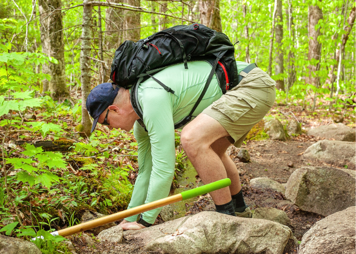 A man wearing a hiking pack, shorts, a long-sleeve green shirt, and a baseball cap leans down to grab something