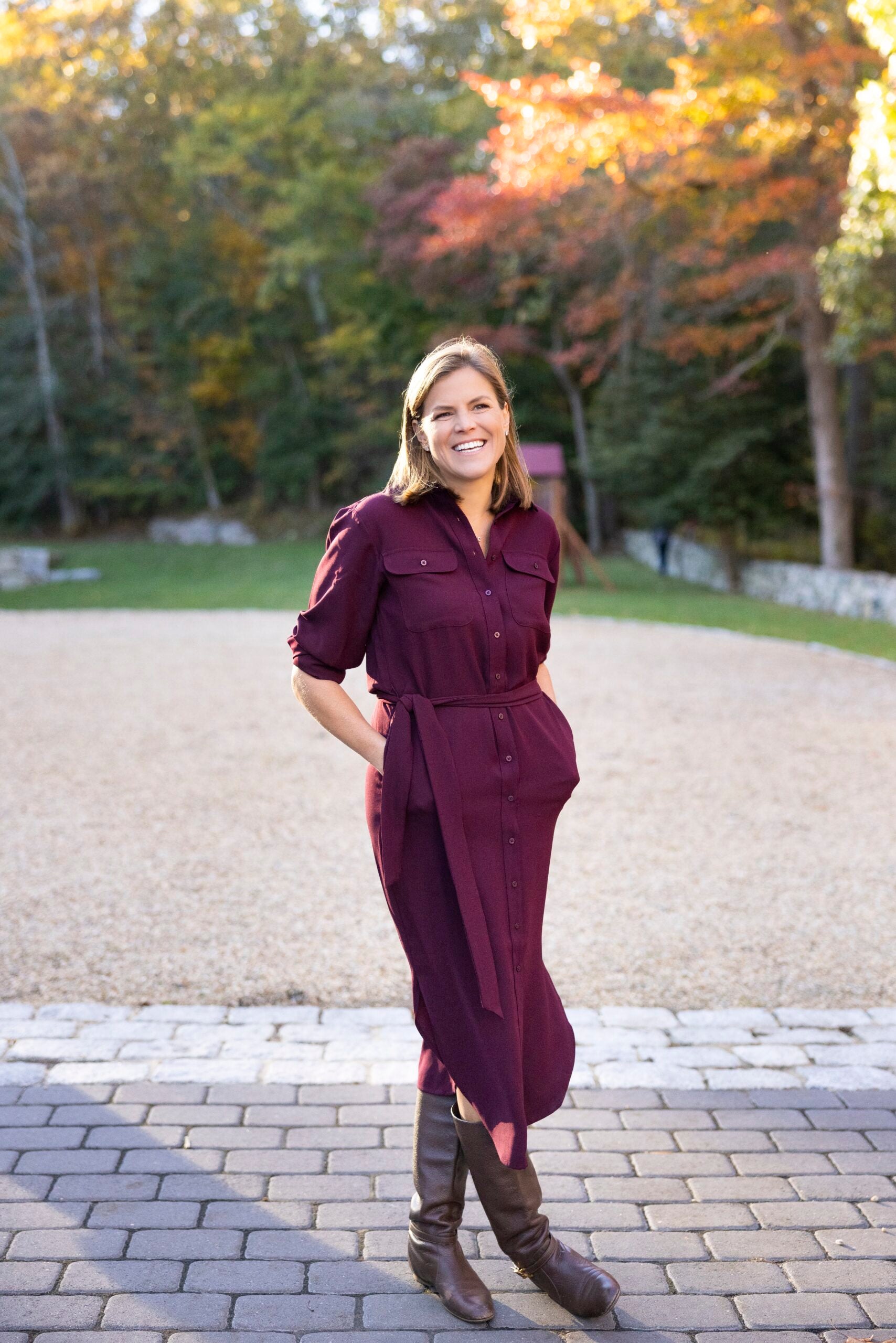 Cornell stands in front of fall foliage. She wears a long burgundy dress and leather boots.