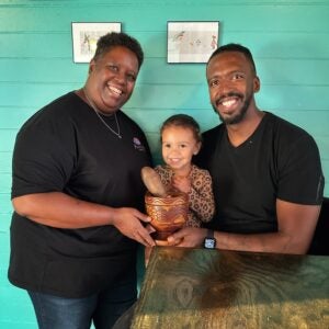 A Black man and woman smile at the camera with a young Black girl in between them while holding a wooden cup in front of them